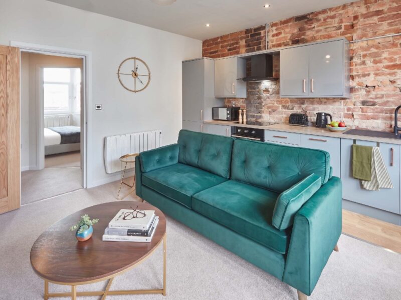 Living area with teal sofa and modern kitchen at Umpires Watch, Cricket View.